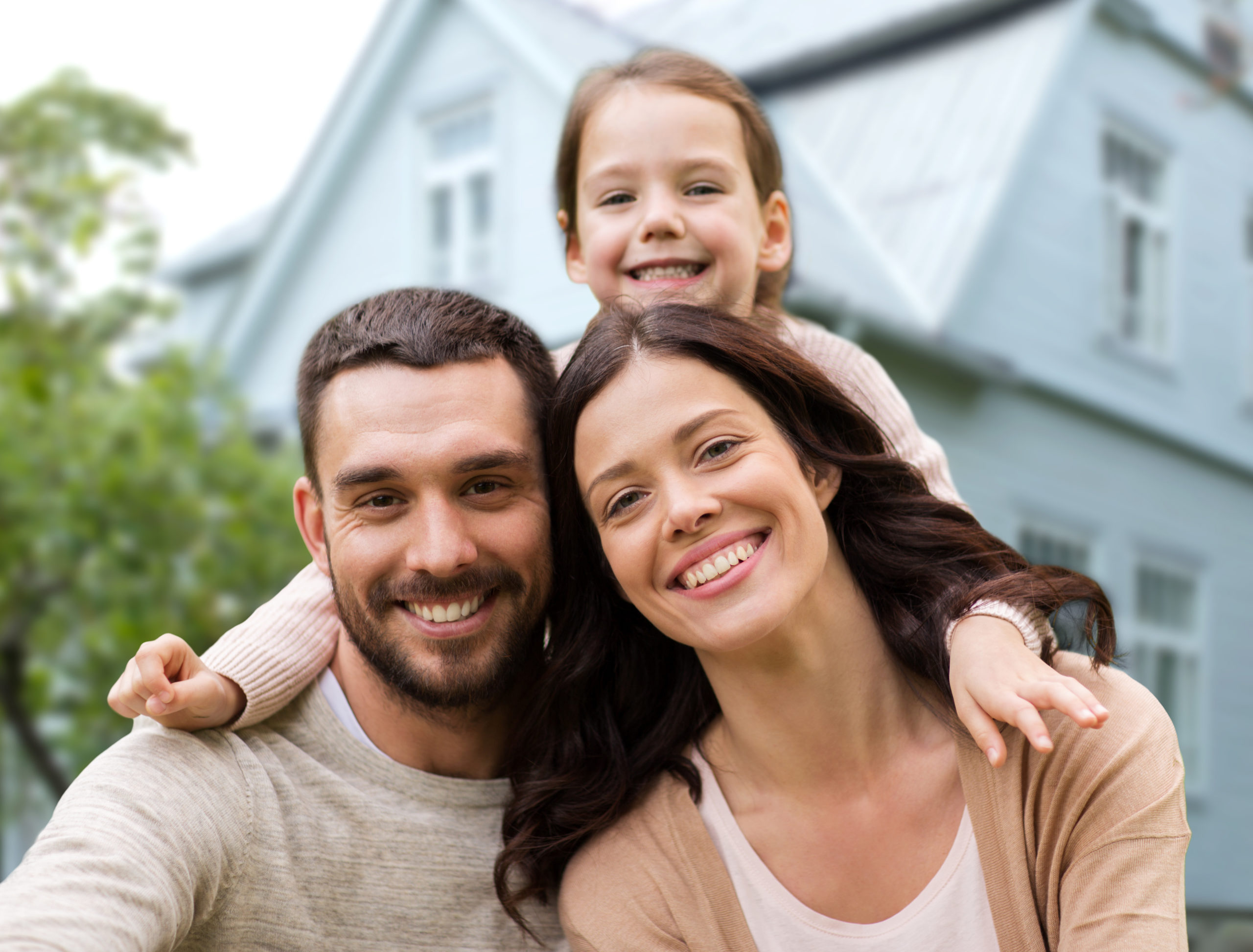 Glückliche Familie mit Kind auf Schultern vor Haus, alle lächelnd.